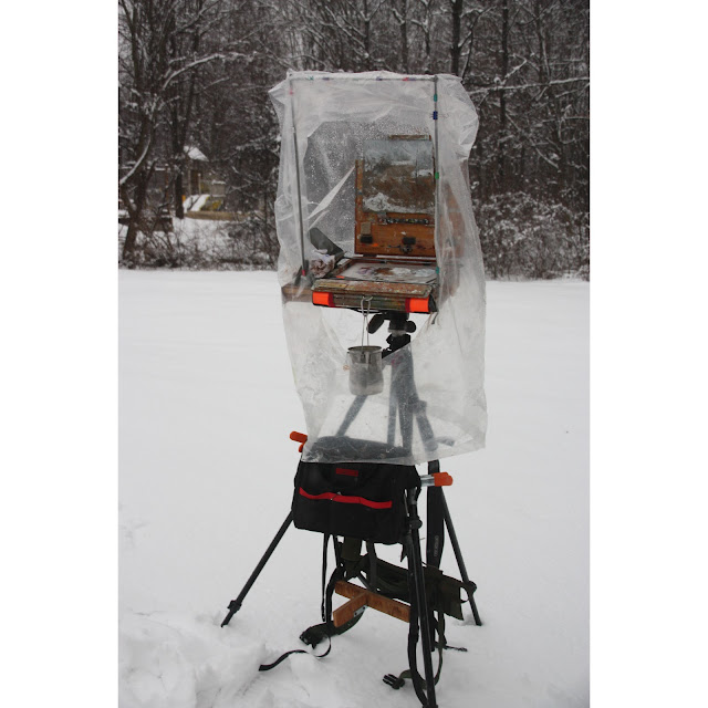 pochade box mounted on tripod in snow, covered by frame with plastic sheeting to protect it from the elements.