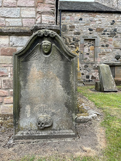 A photo of an old gravestone in Duddingston Kirkyard.  At the top of it there is a face with wings around it and at the bottom is carved a skull.  The engraved name of the person who's grave it was has faded away to nearly nothing.  Photo by Kevin Nosferatu for the Skulferatu Project.