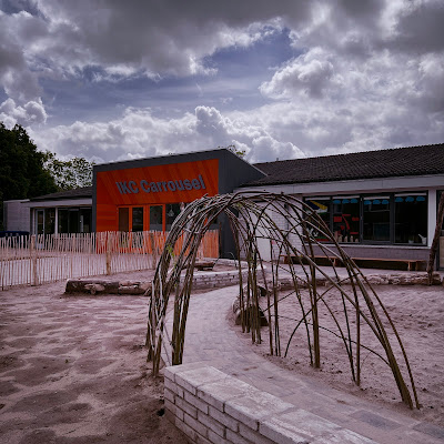Nieuwe vormgeving IKC Carrousel, Zevenaar, mei 2024. Naast de ingang is een 'natuurspeelplaats' te zien met veel zand en takken.