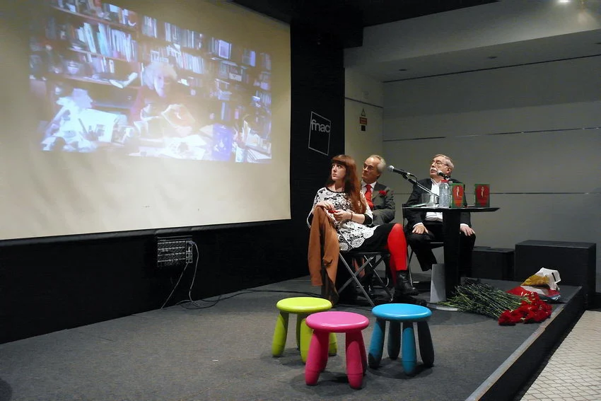 Foto da mesa de apresentação com um écrã por detrás onde se vê a imagem da autora o livro a discursar
