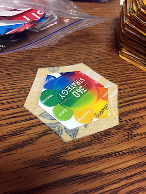 A paper hexagon with a rainbow color wheel diagram, centered on a face-down yellow fabric hexagon, with a pin pushed through the center of the hexagons to hold them together. A small ziploc bag of colorful paper hexagons and a tall stack of yellow fabric hexagons are visible in the background.