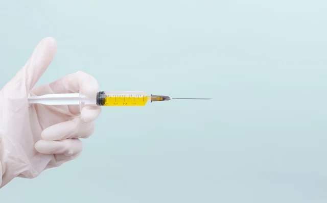 A syringe, filled with a yellow liquid, is held horizontally by a gloved hand emerging from the left side of the image. The scene is set against a light blue background.