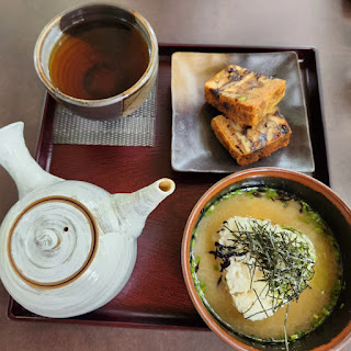 japanese tea service tray. A small cup of tea in left upper corner, with white tea pot in lower left corner. Small slice of nut bread in the upper right corner and a small bowl of miso soup with rice in lower right corner.