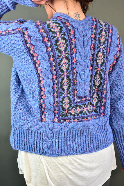 A colorful handknit cardigan viewed from behind. It features stranded colorwork between cables that run a number of directions. The inventive cardigan rests on a deep ribbing. The cables and ribbing are a bright blue, while the colorwork sections show pops of pink with a deep lavender, light gray and dark green.