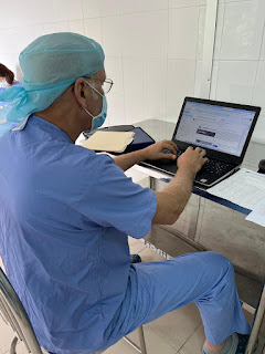 Man wearing scrubs, cap, and mask sitting at table working on a laptop.