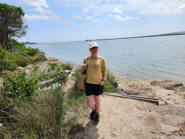 Image of middle aged white woman in yellow & white striped t-shirt and black shorts standing on a small beach