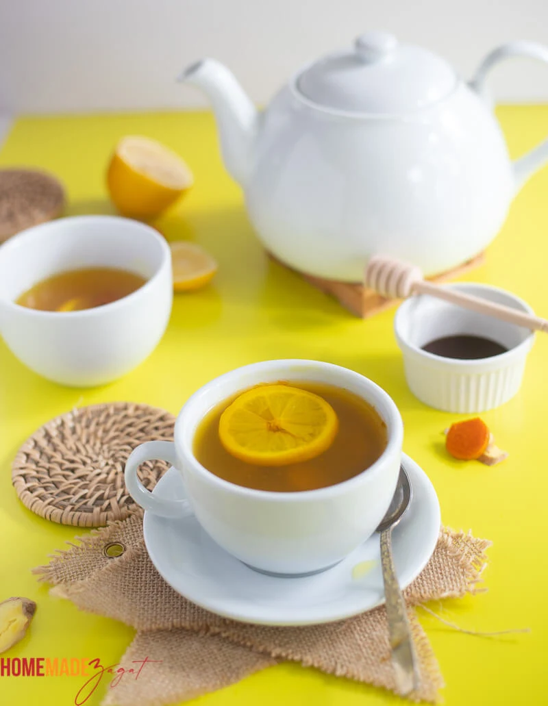 Two white cups of tumeric ginger lemon tea, with honey and lemon on a yellow backdrop