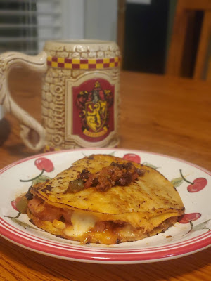 Gryffindor beer stein behind a red and white plate painted with cherries. on top of the plate is a tortilla sandwich with cheese spilling out and a dollop of salsa on top