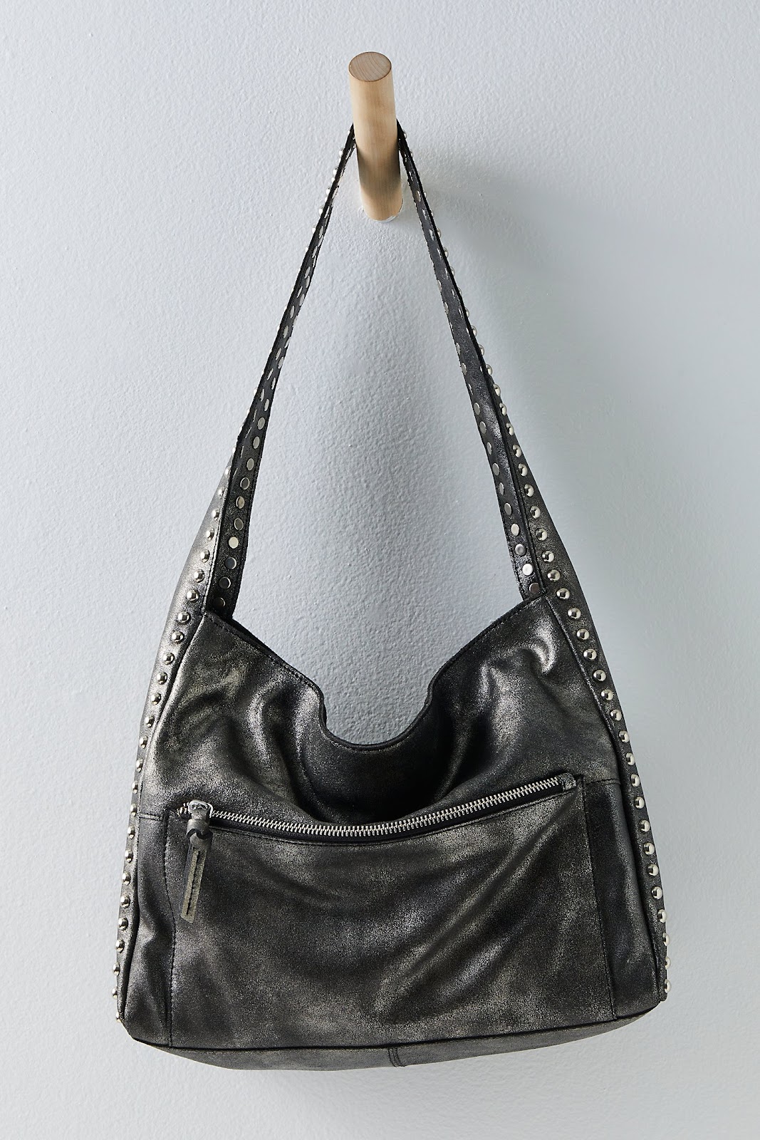 Metallic black leather hobo shoulder bag with thick shoulder strap and silver round studs and silver zipper pocket detail hanging on a wood peg on a white wall