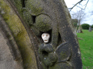A photo of a small ceramic skull (Skulferatu 95) sitting in the decoration at the top of the faded and crumbling gravestone in the Southern Necropolis.  Photograph by Kevin Nosferatu for the Skulferatu Project.
