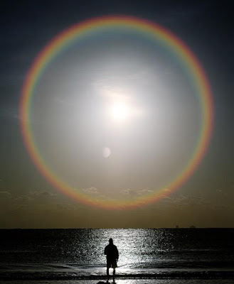 A mysterious fibs trader standing at the ocean shore line with his back turned looking uop at huge sun with a huge holo ring radiating from it.