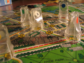 The five clear, colourless plastic pawns from the Scotland Yard board game, on the game board. The board is a stylised map of London, with various street intersections labelled with a number. These numbers are connected by one or more lines of green, yellow, and/or red. The pawns each have a coloured dot to indicate which player's they are, except the one in the centre, belonging to Mr X, which has a white dot marked with a black X.