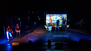 A photograph of the Artrageous performance, showing two singers singing songs on a stage while three artists paint a rendition of Starry Night under black lights.