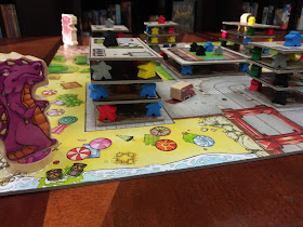 The board at the beginning of the game. A purple lizard monster stands on the corner of the board with a red monster visible on the far corner. The board is made to look like a bird's-eye view of a city; some of the spaces on this board are areas for buildings. These spaces have a meeple in each corner, with a cardboard rectangle on top of them. Four more meeples are on the corner of this rectangle, with another rectangle on top of them. This is topped by a third layer of four meeples and a rectangle; a single meeple stands in the centre of this top rectangle.