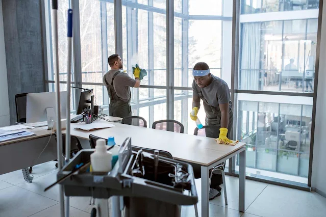 Two cleaners Manchester in uniform clean an office with large windows. One wipes a desk, while the other cleans a window. Cleaning supplies are on a cart in the foreground.