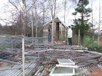 View over garage roof. Timothy stands on outside platform with his back to the camera, looking into the treehouse. Simon, visible inside the treehouse through the gappy wall, swings an axe.