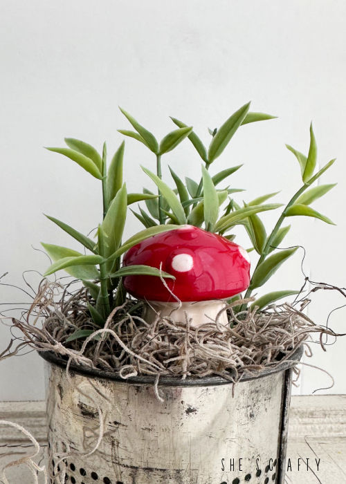 Ceramic red and white mushroom in metal container with greenery.