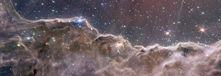 NGC 3324 - A composite image of the Cosmic Cliffs in the Carina Nebula, created with the Webb telescope’s NIRCam and MIRI instruments. Pinkish brown clouds of gas and dust dominate the foreground of the image, glittering with young stars. Behind the glowing, mountainous clouds, the sky appears navy blue, with shining stars and galaxies.