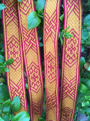 A photograph of several lengths of red and yellow tablet woven band, patterned with floral motifs and lattice work, against a leafy backdrop