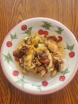 overhead photo of  a wooden table with white plate decorated with painted cherries. on the plate is crumbled pancakes with cinnamon apples, with syrup poured over the top