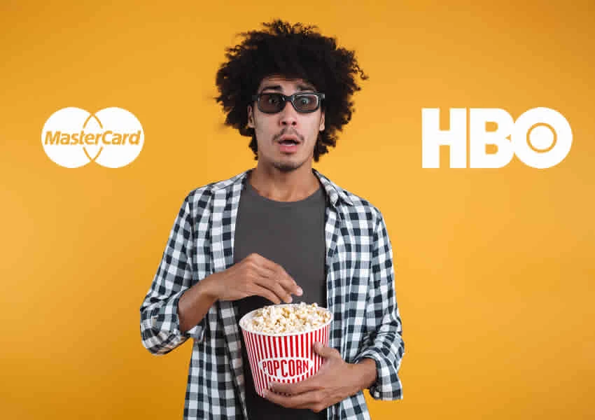 Imagem de fundo amarelo mostra um homem segurando um pote de pipocas abismado olhando para frente ele esta vestindo camisa xadrez e usando oculus 3D de Cinema na imagem também mostra as logos da Mastercard e HBO uma de cada lado do homem.