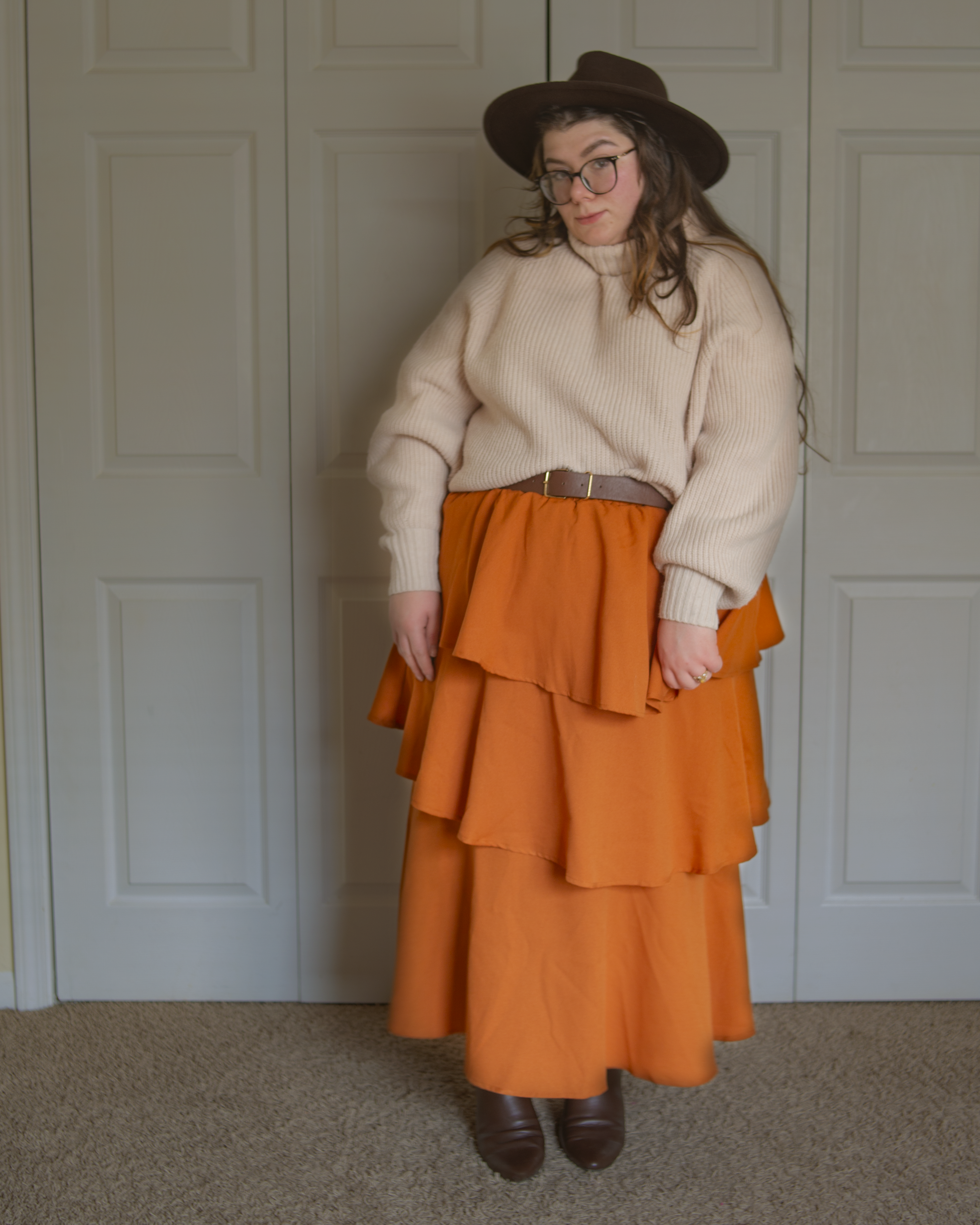 An outfit consisting of a cream sweater tucked into a burnt orange three asymmetrical tiered maxi skirt and brown midi boots.