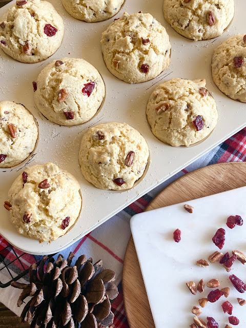 A white muffin tin of cranberry egg nog muffins with cranberries, pine cones and pecans scattered around.