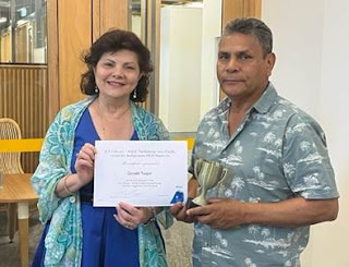 A woman of Greek decent in a blue dress and a colourful scarf poses next to a man of Aboriginal Australian decent. She is passing him a certificate, and he holds a small trophy.