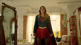 A screenshot of Supergirl, a young, blonde white woman in a dress with a blue bodice, short red skirt, and red cape, standing in her apartment. The place is a converted warehouse with red brick walls and floor to ceiling windows made up of many smaller leaded panes and covered by gauzy white drapes.