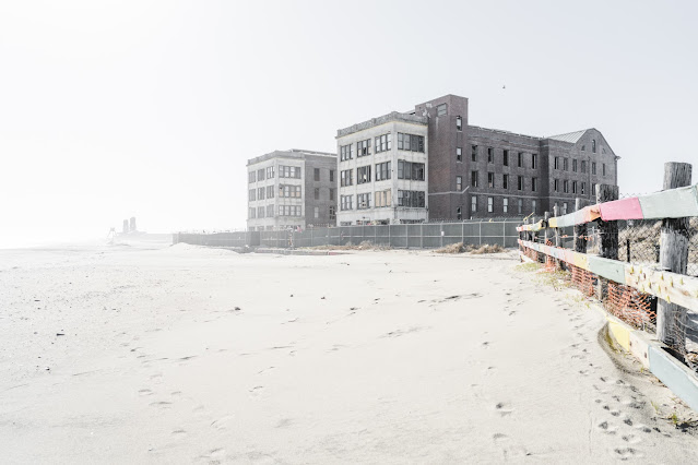 An overexposed photograph of a deserted beachfront with the remnants of Neponsit Beach Hospital buildings. The stark, weathered structures stand in contrast to the soft, bright sand of the beach, and a hazy sky suggests a misty or foggy day. Footprints are visible in the sand, leading towards the dilapidated buildings, while a worn wooden fence with sporadic colorful patches lines the border between the beach and the hospital grounds.