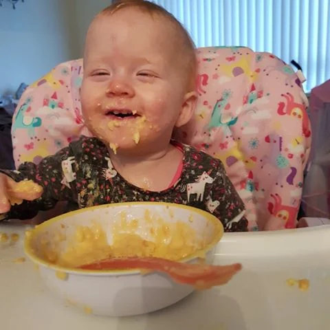 A baby covered in food with a bowl and spoon in front of her