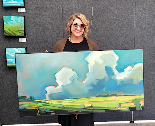 A photograph of oil painter Danielle Clouse Gast holding one of her large paintings. It is a geometric style landscape, with green fields, blue sky and large white clouds.