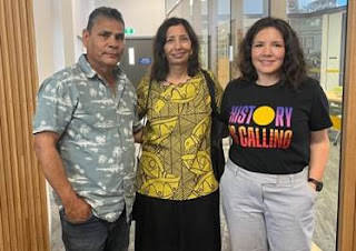 A man and two women stand side-by-side next to a window, smiling at the camera. One of the women is wearing a shirt that says "history is calling" in colours and designs reminiscent of the Australian Aboriginal flag.