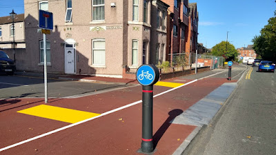 A side road junction which has a raised area surfaced red over which a shared pavement and cycle track crosses. A white line to split the space has been added as well as a pair of rectangular yellow areas to represent tactile paving.