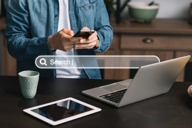 Um homem usando camiseta branca e camisa jeans por cima usa um celular. Na frente da tela, há a ilustração de uma barra de pesquisa escrito "search".