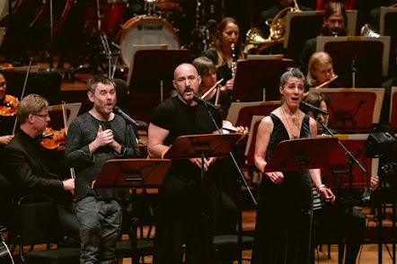 Photo. Three singers, two men in informal clothing and one woman in a gown, standing in front of an orchestra, reading music from music stands and singing.