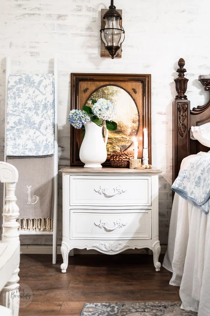 white brick wall, ladder with blankets, nightstand with hydrangeas in ironstone pitcher, candlesticks and rustic pumpkin