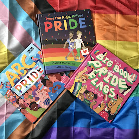 A photo of three Pride picture books on a large Progress Pride Flag, At the top-center is 'Twas the Night Before Pride by Joanna McClintick and Juana Medina. ABC Pride by Dr Elly Barnes MBE, Louie Stowell and Amy Phelps in on the bottom left, partially under 'Twas the Night Before Pride. It's diagonal, top left to bottom right. The Big Book of Pride Flags by Jessica Kinglsye Books and Jem Milton is at the bottom right, particially on top of 'Twas the Night Before Pride. It's diagonal, top right to bottom left.