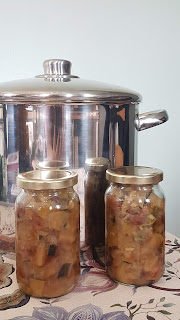 Two jars at the front with chutney inside on a floral table cloth. Behind them is a large, shiny, saucepan
