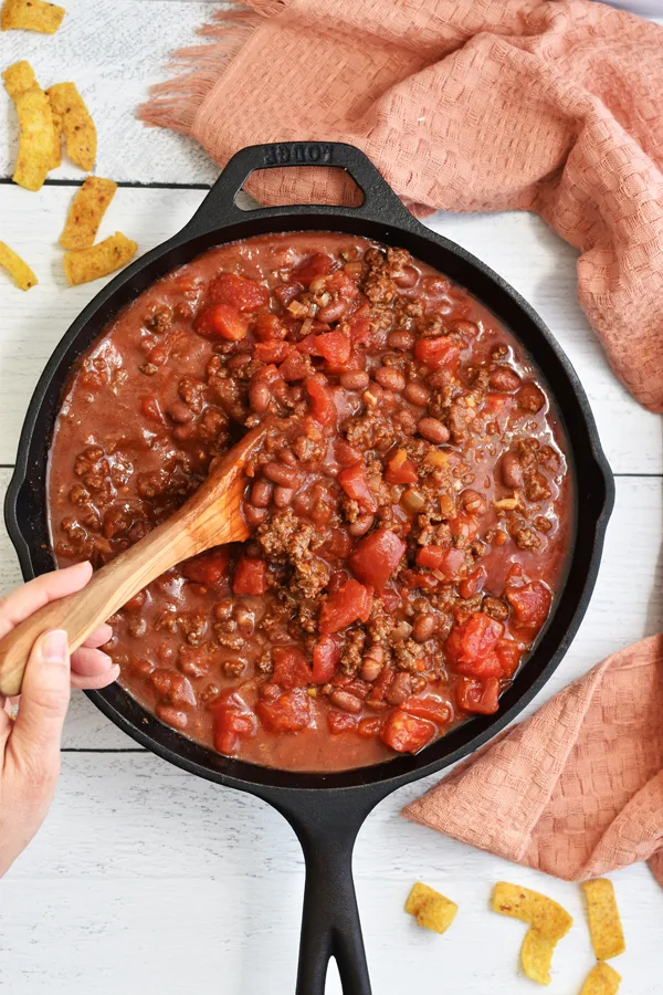 A hand holding a wooden spoon stirring ingredients into browned ground beef in a cast iron skillet, creating a flavorful and well-blended mixture.