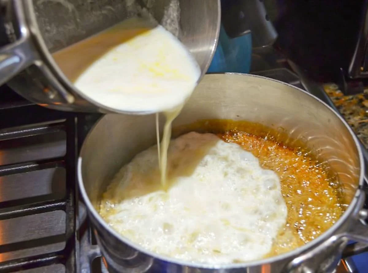 Hot cream and butter being poured into Caramelized Sugar in a stainless steel pot.