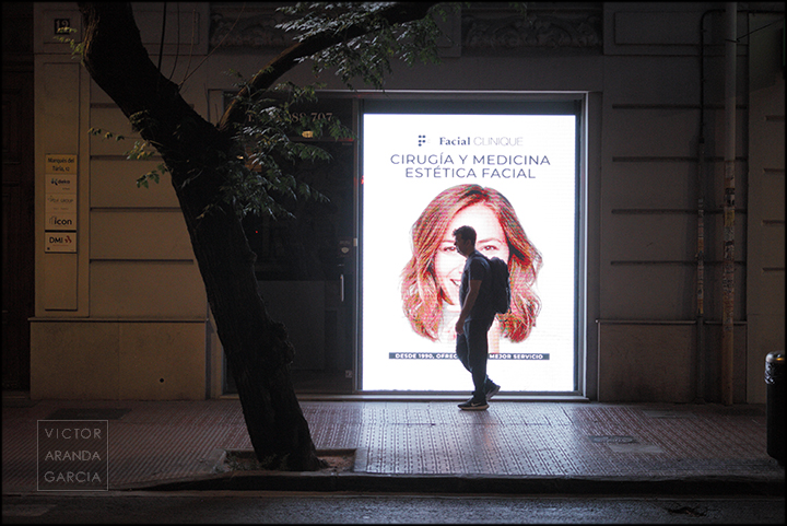 Fotografía de una calle de Valencia de noche con un cartel publicitario y un viandante pasando delante