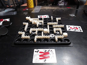 The board during a game in progress. Several barriers shaped like a row of trees are placed between two adjacent spaces to form a maze around the board. Several of the 'barrels' can be seen around the board. There are also rock tokens in a few places. Cards denoting the cardinal directions (north, south, east, and west) are placed around the board to help people remember which way is which.