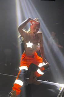 House of Happiness male sober club dancer wearing orange workman's hi-vis leggings while dancing in a style of martial arts movements with a female hairstyle beyond shoulder length