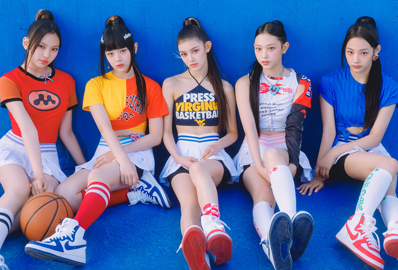 A shot of NewJeans [From Left to Right: Hyein, Hanni, Danielle, Haerin, Minji] on a basketball, wearing T-shirts, skirts and Nike hi-tops. This is one of the group looks from the music video for “Super Shy”.