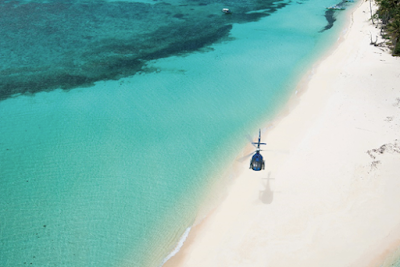 A helicopter flying over a white-sand beach next to a turquoise sea.