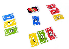 na zdjęciu rozgrywka trzyosobowa, na środku leży stos kart zakrytych, po bokach po trzy odkryte karty graczy i wyłożona startowa karta o wartości pięć