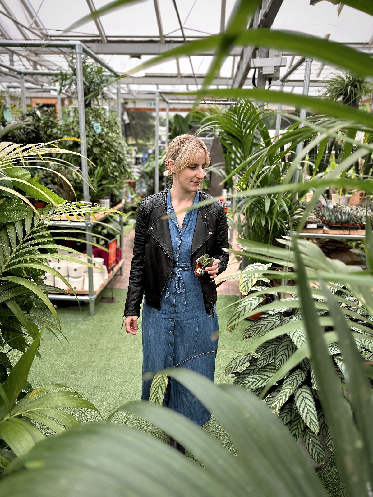 Amy is in a garden centre surrounded by plants; wearing a denim maxi dress and leather jacket