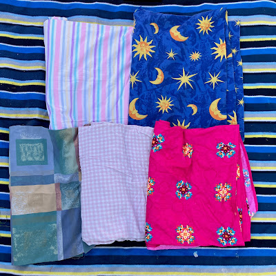 Five pieces of fabric have been partially unrolled to show off their patterns. The top left has pastel blue, purple, yellow, pink, and white stripes. Top right is dark blue with suns, moons and stars. Bottom left simulates block printing with large squares and rectangles of greens, blue and yellow. Bottom middle is a pink and white gingham with half inch squares. Bottom left has a deep pink background, with equally spaced mandala-like flower patterns worked in yellow, orange, maroon, teal, and sky blue.