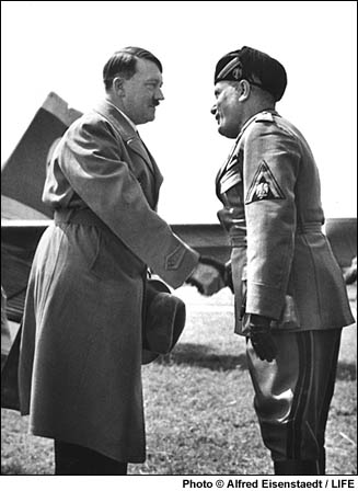 Imagem em preto e branco de Adolf Hitler apertando a mão de Benito Mussolini. Mussolini veste roupas militares e Hitler está de casaco. O ambiente é um gramado e os dois estão à frente daquilo que parece ser um avião.